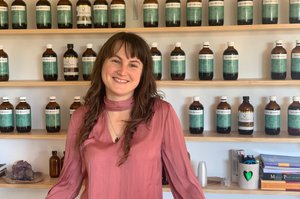 brunette woman with fringe smiles at camera with rows of herbal medicine stocked behind her